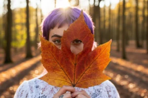 Modella con capelli viola che regge una grande foglia d'autunno arancione davanti al viso, bosco dorato