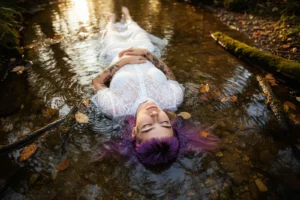Modella con capelli viola sdraiata nel ruscello, abito bianco in pizzo, occhi chiusi