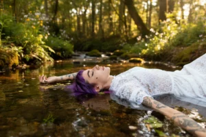 Modella con capelli viola sdraiata nel ruscello con braccia aperte, abito bianco, luce naturale