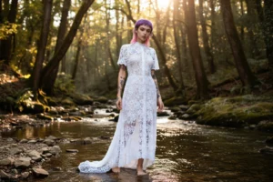 Modella con capelli viola e abito bianco in pizzo in un ruscello nel bosco, luce dorata del tramonto