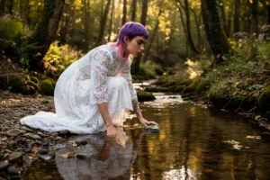 Modella inginocchiata sul bordo del ruscello che tocca l'acqua con la mano, abito bianco