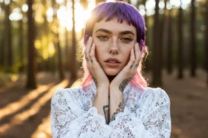 Primo piano modella con mani che incorniciano il viso, luce calda di pineta, piercing e tatuaggi