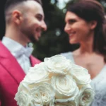 Bouquet di rose bianche in primo piano, sposi sorridenti sullo sfondo, sposo in abito rosso | Fabio Zenoardo