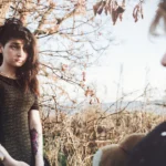 Modella appoggiata al tronco in primo piano, altra figura sfocata in secondo, fotografia moda atmosferica