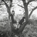 Fotografia in bianco e nero, due ragazze sedute nella biforcazione di un albero, paesaggio autunnale