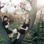 Due ragazze tra pi&ugrave; alberi, una sulla biforcazione del tronco, posa dinamica, fotografia moda outdoor