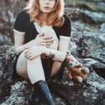 Modella seduta su rocce ricoperte di muschio, tiene un orsacchiotto, espressione pensierosa, fotografia moda