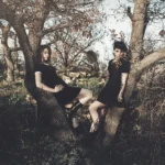 Due ragazze sedute su rami di un albero, spalle voltate in direzioni opposte, fotografia fashion moda