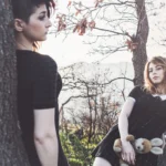 Ragazza in primo piano appoggiata ad albero guarda l'altra, tatuaggio sul braccio visibile, fashion photography