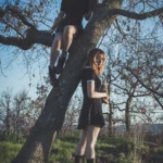 Una ragazza arrampicata sull'albero e una sotto, abiti neri, cielo blu, fotografia fashion outdoor