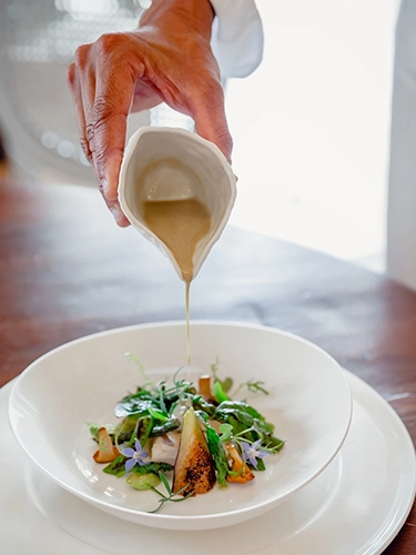 Home Mano di chef che versa salsa dorata su piatto gourmet con verdure e fiori eduli