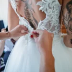 Mani allacciano l'abito da sposa in pizzo trasparente, tatuaggi visibili sulla schiena | Fabio Zenoardo