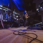 Musicista con chitarra acustica ripreso dal basso sul palco, luci blu e strumentazione live