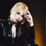 Ritratto di Yuriko Tiger in latex nero con luce bassa, capelli biondi corti, espressione decisa, fotografia cosplay