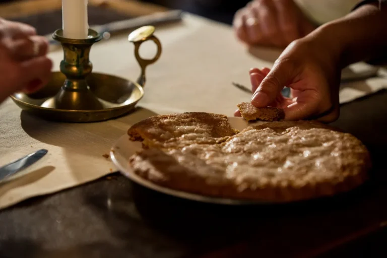 Mani che spezzano una fetta di torta su piatto bianco, candeliere in ottone, atmosfera calda e rustica