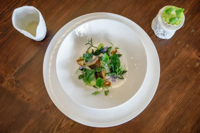 Vista dall alto di filetto di pesce con erbe, verdure di stagione e fiori viola su piatto bianco, bricco e coppetta a lato