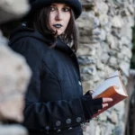 Attrice Ghost Tour in cappotto nero e cappello con libro, makeup gotico, muri in pietra