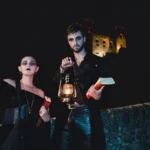 Coppia in costume dark con lanterne e libri, makeup gotico, castello medievale notturno