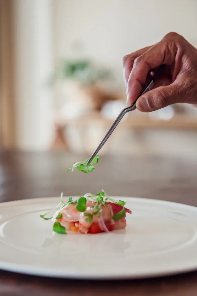 Chef con pinzette posa microgreens su tartare di gamberi su piatto bianco, luce naturale, ristorante Imperia