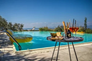 Piscina infinity con tavolo aperitivo in primo piano, sdraio, olivi e vista lago mare all orizzonte al tramonto