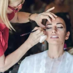 Make-up artist con occhiali rossi applica trucco alla sposa in vestaglia bianca | Fabio Zenoardo