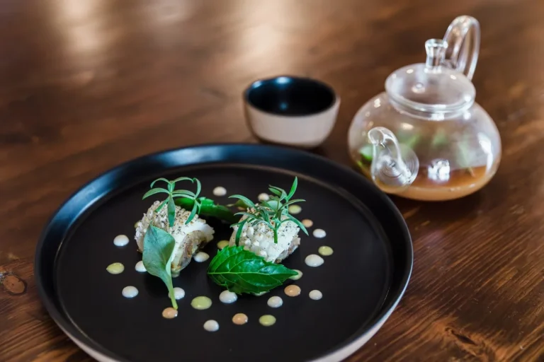 Due porzioni di pesce in crosta di sesamo ed erbe su piatto rotondo nero con gocce di salsa verde, teiera in vetro