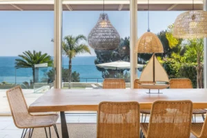Sala da pranzo con lampade in rattan, tavolo in legno e sedie, ampia vetrata con vista panoramica sul mare