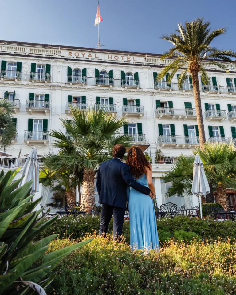 Coppia abbracciata di spalle davanti alla facciata del Royal Hotel Sanremo con palme e giardino