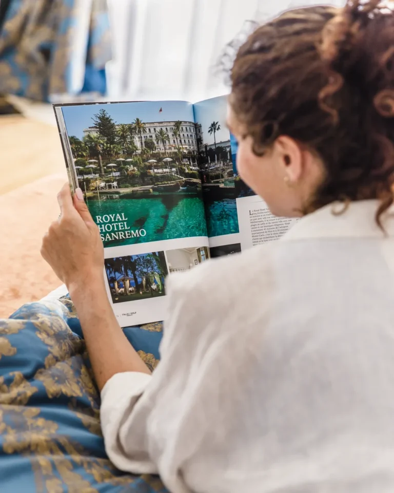 Donna sdraiata sul letto con accappatoio che legge rivista Royal Hotel Sanremo, camera elegante