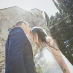Sposi si baciano davanti a una chiesa in pietra con cipressi, sposa con tatuaggi | Fabio Zenoardo