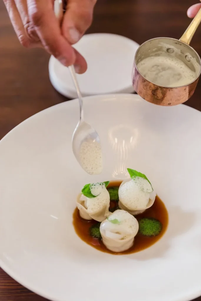 Primo piano di tortellini in brodo scuro con schiuma aromatica versata da cucchiaio, erbe verdi, pentolino rame