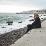 Fotografo fashion Liguria - modella seduta riva giacca nera mare mosso ligure